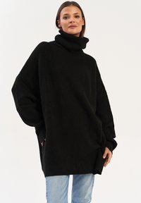 Czarny, oversize sweter z golfem o prążkowanej strukturze, z długimi rękawami i bocznymi kieszeniami, noszony z jasnoniebieskimi dżinsami. Prosty, nowoczesny design.