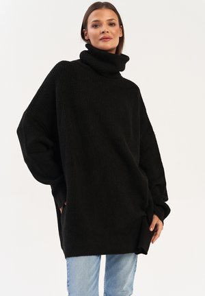Czarny, oversize sweter z golfem o prążkowanej strukturze, z długimi rękawami i bocznymi kieszeniami, noszony z jasnoniebieskimi dżinsami. Prosty, nowoczesny design.
