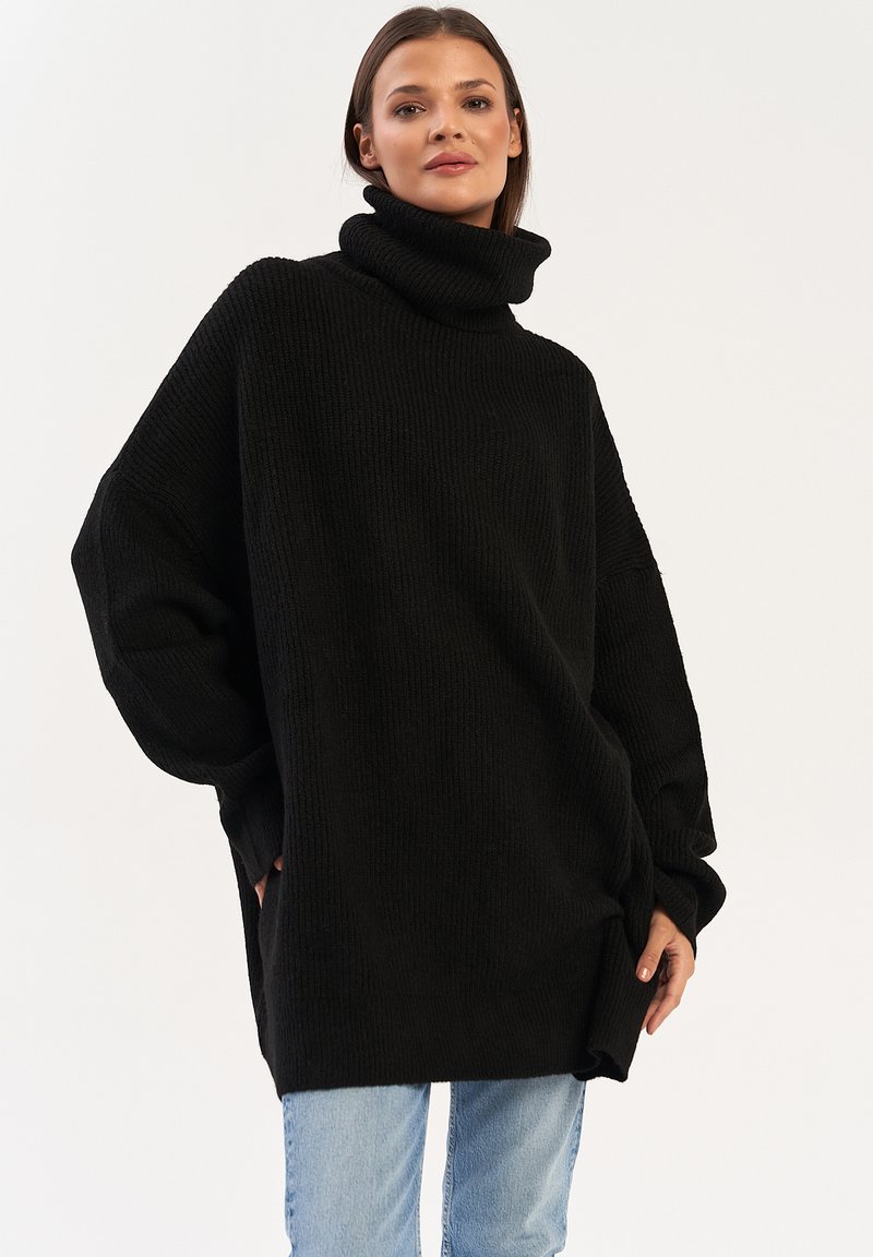 Czarny, oversize sweter z golfem o prążkowanej strukturze, z długimi rękawami i bocznymi kieszeniami, noszony z jasnoniebieskimi dżinsami. Prosty, nowoczesny design.
