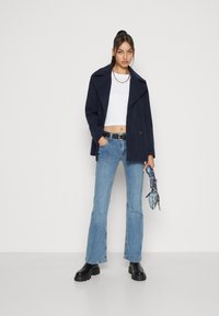 Mörkblå dubbelknäppt kappa, vit croppad topp, ljusblå utsvängda jeans och svarta ankellångstövlar. Accessoar: mönstrad blå handväska.