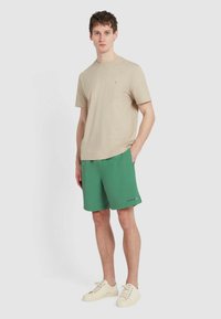 T-shirt beige a maniche corte con collo rotondo, abbinato a pantaloni da bagno verdi. I pantaloni presentano un dettaglio con logo e sono di lunghezza sopra il ginocchio.