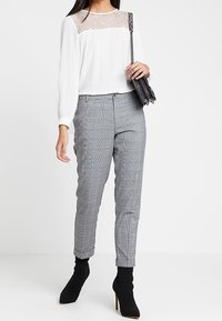 Blusa bianca con dettagli in pizzo sul colletto, abbinata a pantaloni grigi a quadri. Il modello indossa stivaletti neri e tiene una borsa nera.