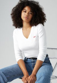 Bijela majica dugih rukava s V-izrezom i malim crvenim Levi's logom, u kombinaciji s plavim trapericama visokog struka. Glatka tekstura, ležeran kroj.