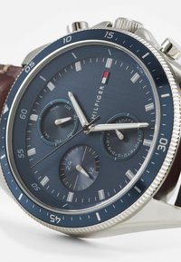 Reloj cronógrafo de esfera azul con caja plateada, correa de cuero marrón y marcadores de fecha. Presenta tres subesferas y acentos pulidos.