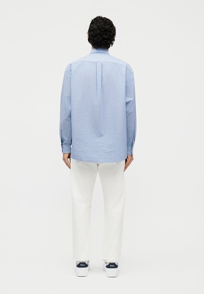 Άνδρας με σγουρά μαλλιά που φοράει ανοιχτό μπλε πουκάμισο με μακριά μανίκια, λευκό παντελόνι και λευκά αθλητικά παπούτσια, κοιτάζοντας προς τα πίσω σε ένα απλό φόντο.