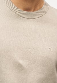 Κοντινό πλάνο ενός ατόμου που φοράει μπεζ πουλόβερ με ριμπ ύφανση και στρογγυλή λαιμόκοψη, με ένα μικρό κεντημένο λογότυπο στο στήθος.