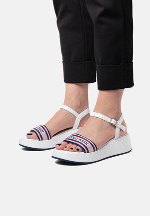 White platform sandals feature a wide strap with a blue and red logo. The straps are adjustable with a silver buckle.