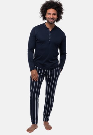 Mann mit lockigem Haar, der ein marineblaues Langarmshirt und marineblaue Hosen mit weißen vertikalen Streifen trägt, steht barfuß und lächelt.