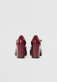 Chaussures à talon bloc en cuir bordeaux avec une finition brillante, dotées d'un bout arrondi et d'un léger accent de boucle sur le côté.