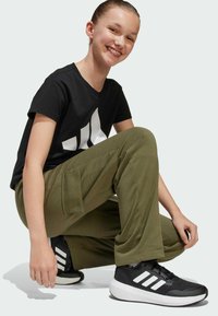 T-shirt nera con logo bianco, pantaloni verde oliva con tasca laterale e sneakers nere con dettagli bianchi, presentati in una posizione accovacciata.