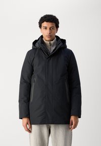 Chaqueta negra impermeable con capucha, bolsillos con cremallera y una textura suave. Llevada sobre un suéter ligero acanalado, con un diseño de corte recto.