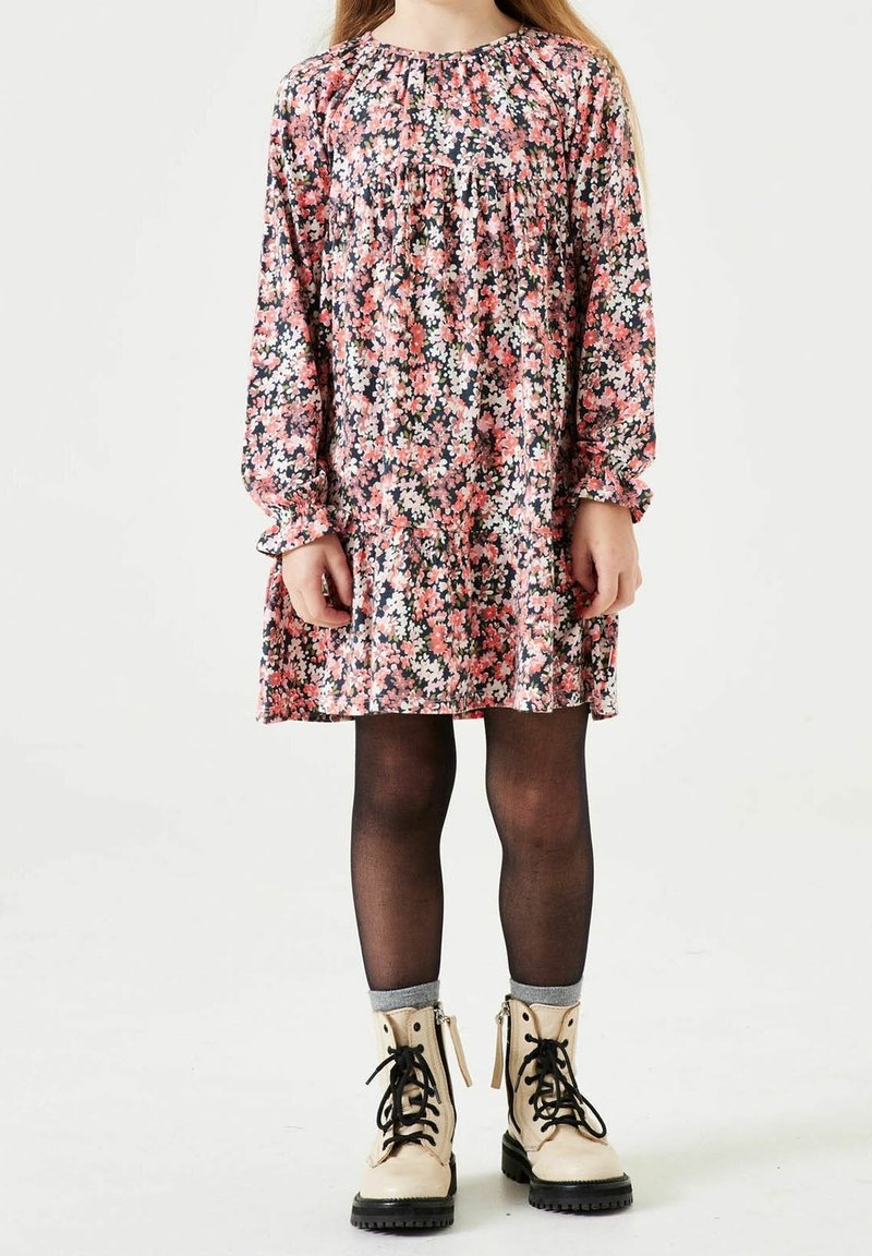 Fille portant une robe fleurie à manches longues, des collants noirs et des bottines beiges à lacets, debout devant un fond blanc.