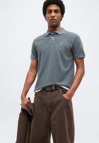 Άνδρας που φορά γκρι πόλο μπλούζα και καφέ παντελόνι, κρατώντας καφέ σακάκι στο αριστερό χέρι, στέκεται μπροστά σε απλό φόντο.