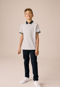 Szürke galléros póló fekete gallérral és csíkkal, tengerészkék nadrággal és fehér tornacipővel párosítva. Rövid ujjú, texturált anyaggal.