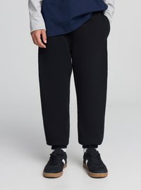 Pantaloni in pile nero con polsini a coste, caratterizzati da una vestibilità comoda. Abbinati a sneakers nere con una striscia bianca e suola in gomma color beige.