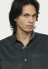 Chemise en velours côtelé vert foncé avec une texture à côtes verticales, des boutons-pression en argent, un col boutonné et une poche sur la poitrine.