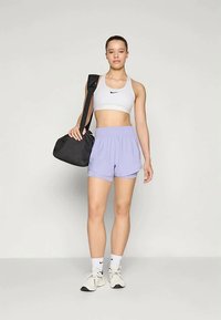Sportliches Outfit mit einem weißen Sport-BH, lilafarbenen Shorts und weißen Sneakers. Schwarze Tasche mit Schulterriemen. Glatte Stofftexturen.