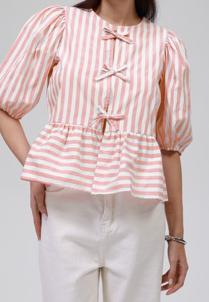 Kvinde iført lyserød og hvidt vertikalt stribet bluse med tre sløjfer ned foran og puffede ærmer, sat sammen med hvide bukser.