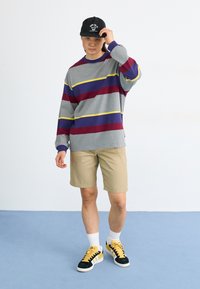 Chemise grise à manches longues avec des rayures horizontales rouges, violettes et jaunes, shorts kaki, et casquette noire; baskets jaunes et noires.