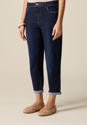 Frau trägt dunkelblaue hochgekrempelte Jeans und beige Mokassinschuhe, mit einem pinken Armreif am linken Handgelenk vor einfachem Hintergrund.