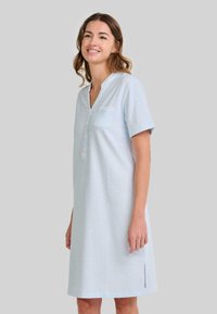 Mujer sonriendo, vistiendo un vestido de manga corta hasta la rodilla de color azul claro con botones en el frente, bolsillo en el pecho y abertura lateral, contra un fondo liso.