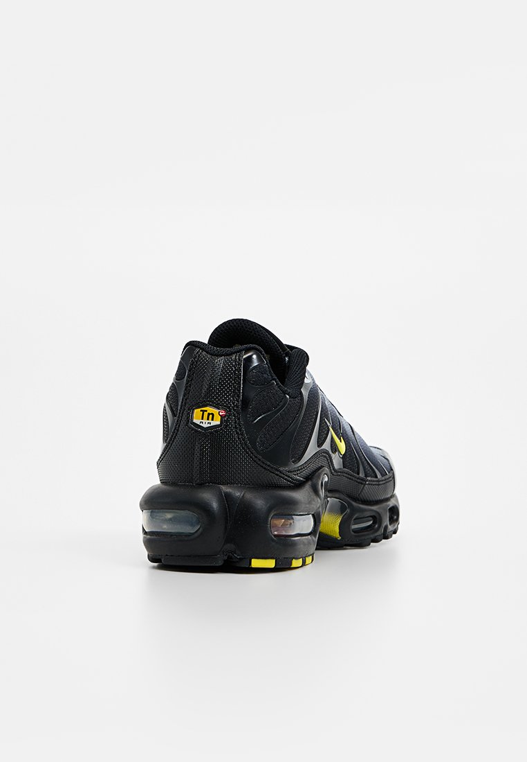 Chaussure de sport noire avec une tige en maille, accents jaunes et amorti à l'air dans la semelle. Caractérisée par un marquage distinctif sur le talon.