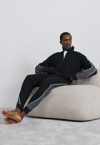 Čierna bunda na zips so sivými detailmi a zodpovedajúce nohavice s bielym lemovaním. Materiál pôsobí mäkko s voľným strihom. Modelka sediaca naboso.