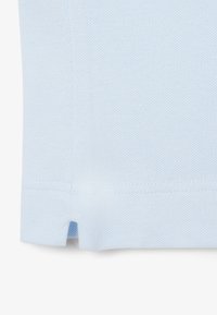 Vaaleansininen poolopaita, joka on valmistettu teksturoidusta kangasmateriaalista. Paidassa on sivuhalkiot helmassa, ja sen muotoilu on puhdas ja yksinkertainen ilman näkyviä logoja tai koristeita.