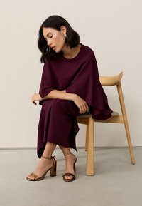 Bordó aszimmetrikus ruha széles ujjakkal, fa széken ülve viselve. Aranyszínű pántos magas sarkú cipő texturált pánttal. Egyszerű háttér.