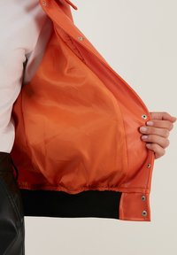Oranje leren jack met een gladde textuur en een elastische zwarte zoom, voorzien van zilveren drukknoopjes en een oranje voering.