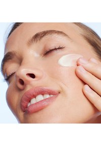 Crema bianca applicata sulla pelle della guancia. Inquadratura ravvicinata di un volto, che mostra una carnagione liscia, ciglia castano chiaro e labbra rosa tenue. Sfondo luminoso.