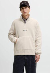 Young man wearing a beige JJ Studios fleece with front pocket, checkered shirt underneath, black jeans, and a black cap labeled "Originals."