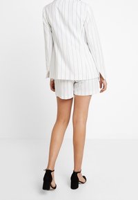 Valkoinen raidoitettu bleiseri ja siihen sopivat shortsit, joissa on muotoiltu malli, yhdistettynä mustiin korkokenkiin. Siisti kangas ja rakenteellinen muotoilu.