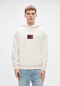 Νέος άνδρας φορώντας μια λευκή κουκούλα με κόκκινο-μαύρο λογότυπο "HUGO" και ανοιχτόχρωμα τζιν, στέκεται μπροστά σε έναν λευκό φόντο.