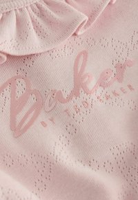 Tessuto lavorato a maglia rosa tenue con piccoli fori disposti a motivo floreale e la parola "Baker" stampata con grandi lettere corsive.