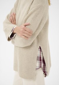Maglione beige lavorato a maglia con un taglio morbido e fessure laterali, sovrapposto a una camicia a quadri bianca e bordeaux, caratterizzato da una texture a coste e maniche larghe.