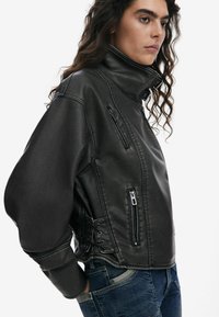 Black leather jacket with a high collar, featuring zippered pockets and lace-up detailing on the sides. Worn over denim jeans.