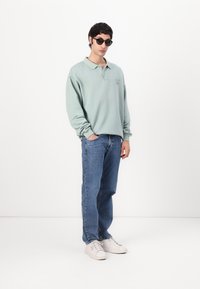 Green cotton polo shirt with long sleeves, blue denim jeans, and white sneakers. Model wears black sunglasses, standing against a white background.