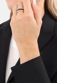 Anelli dorati impilati su una mano, con bande scure e chiare alternate. La mano è parzialmente coperta da un guanto in tessuto nero.