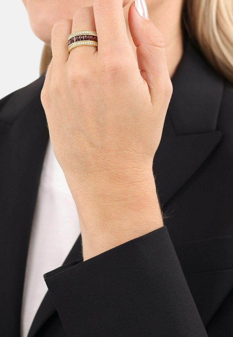 Anelli dorati impilati su una mano, con bande scure e chiare alternate. La mano è parzialmente coperta da un guanto in tessuto nero.