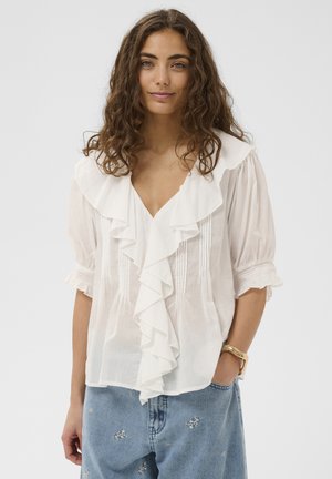 Žena s kučeravými hnedými vlasmi nosí bielu blúzku s volánikmi a nafúknutými rukávmi a svetlomodré rifle s drobnými vyšívanými kvetmi.