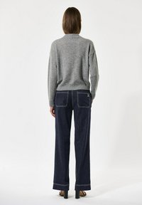 Persona ģērbjas pelēkā garajā piedurknē un tumši zilās platu kāju džinsos, stāvot ar rokām blakus, vērsta prom no kameras.