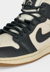 Κοντινό πλάνο ενός μαύρου και εκρού Air Jordan sneaker με υφασμένο μαύρο λογότυπο Nike και σκούρες κορδόνια σε ανοιχτή ταμπά σόλα.