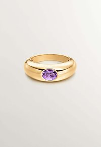 Anillo de tono dorado con un diseño suave y redondeado, y una gema ovalada púrpura en el centro. El acabado pulido resalta las facetas de la gema.