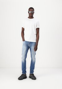 Camiseta de algodón blanca con mangas enrolladas, jeans de mezclilla azul claro y zapatillas deportivas oscuras. Gafas de sol reflectantes completan el look.