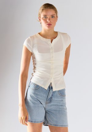 Ung kvinde iført en tætsiddende hvid kortærmet knapbluse og lyseblå højtaljede denimshorts, stående mod en ensfarvet baggrund.