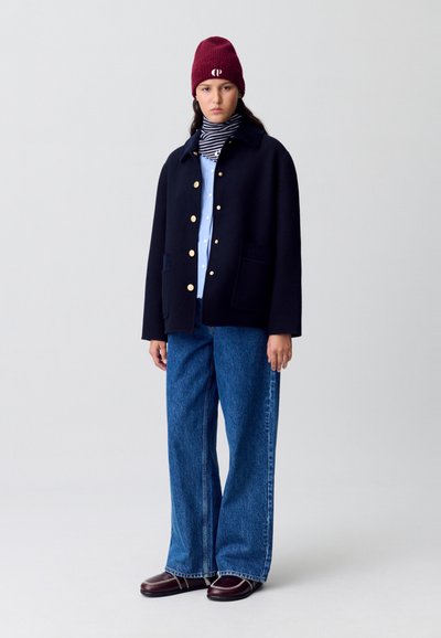 Cappotto in misto lana blu marino con bottoni dorati, indossato sopra una camicia azzurra chiara, abbinato a jeans in denim a gamba larga e a un berretto lavorato in acrilico color bordeaux.