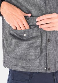 Gilet grigio texture con un primo piano di una mano che si infila in una tasca. Presenta dettagli metallici a scatto e un motivo intrecciato sul tessuto.