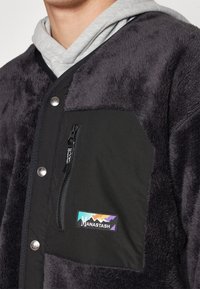 Primo piano di una persona che indossa una giacca in pile grigio scuro con una tasca sul petto nera con cerniera e un'etichetta patch del logo multicolore di Manastash.