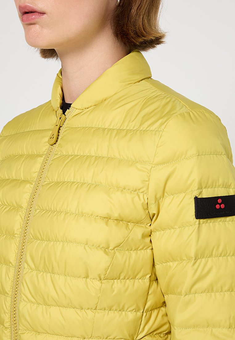 Gelbe Pufferjacke mit vertikalen Nähdetails, Stehkragen, Frontreißverschluss und einem schwarzen Etikett am Ärmel mit roten Punkten.